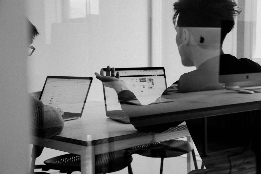 En una sala de reuniones, dos personas están hablando. Tienen ordenadores portátiles sobre la mesa y un muro de cristal refleja las pantallas. La imagen está en blanco y negro.